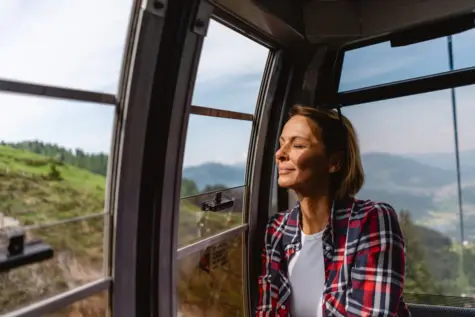 Frau sitzt in einer Bergbahnkabine und blickt lächelnd durch ein Fenster nach draußen.