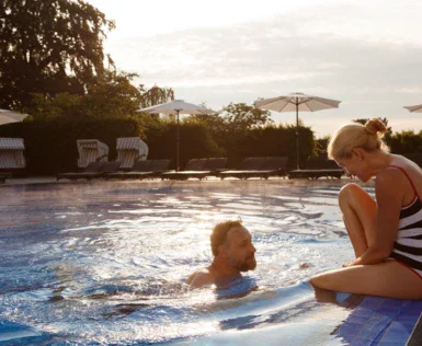 Ein Mann im Pool spricht zu einer Frau, die am Rand sitzt, während die untergehende Sonne den Himmel und das Wasser in warmen Tönen erleuchtet und Liegestühle und Sonnenschirme im Hintergrund auf eine entspannende Atmosphäre hindeuten.