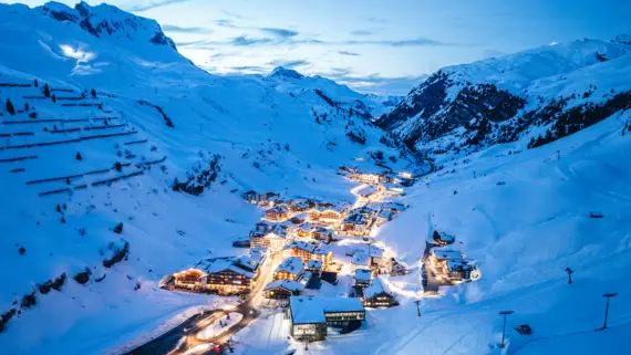 An aerial view of a small village illuminated by light in a valley surrounded by snow-capped mountains. 