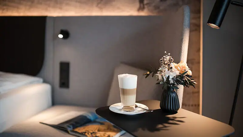 Ein Beistelltisch mit einem Latte Macchiato, einer aufgeschlagenen Zeitschrift und einer Vase mit Blumen neben einem Bett mit gepolstertem Kopfteil vor einer kunstvoll gestalteten Tapete..