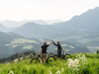 Zwei Radfahrer stehen auf einer grünen Bergwiese in den Kitzbüheler Alpen, geben sich ein High-Five und genießen die atemberaubende Aussicht auf das weite Tal und die umliegenden Berge.