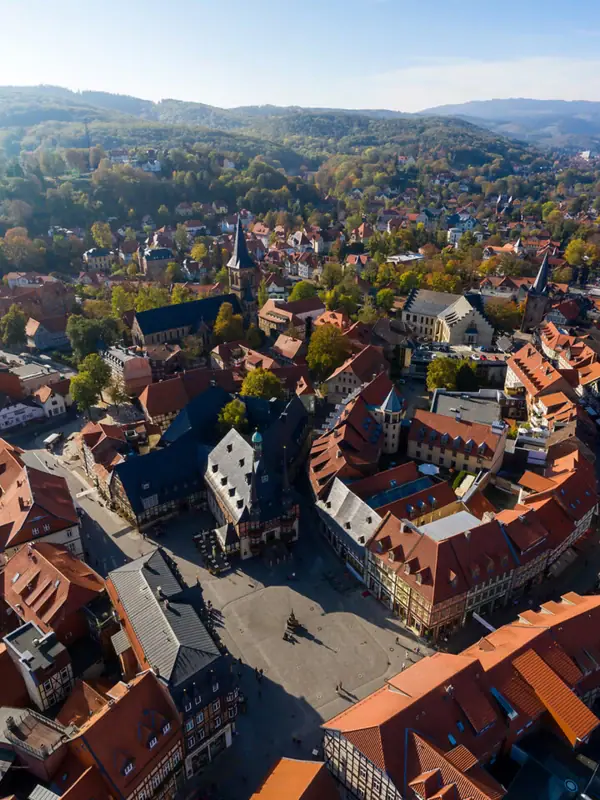Luftaufnahme einer Stadt mit Häusern und Bäumen, umgeben von Bergen und Wolken am Himmel.