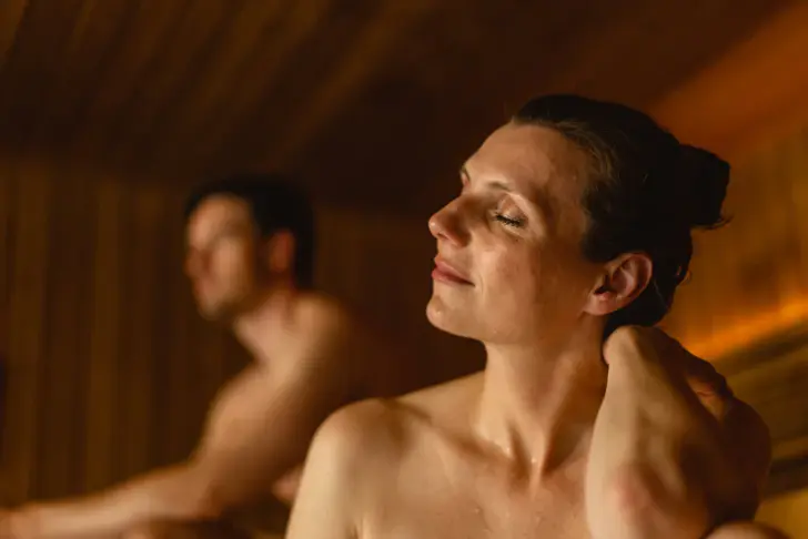 Two people are relaxing in a sauna, the focus is on a woman who has peacefully closed her eyes and slightly tilted her head, visibly enjoying herself. She is in the foreground, while a male person can be seen out of focus in the background. The wooden walls of the sauna and the soft light create a calm and warm atmosphere.
