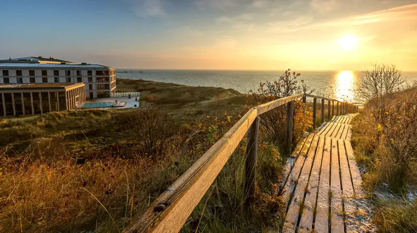 Holzsteg führt zu einem Strand mit Gebäude und Gewässer im Hintergrund.