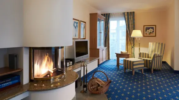 The cozy interior of an upscale hotel room that radiates warmth and comfort. At the center is a modern, rounded fireplace with visible flames behind a shimmering glass front, creating a cozy atmosphere. A woven basket of firewood next to the fireplace enhances the inviting ambience. The furnishings include a wooden cabinet with a flat-screen TV and elegant striped armchairs with matching stools on a blue carpet with floral accents. A window lets in natural light and offers a view outside.