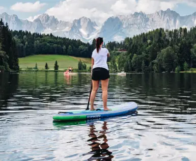 Eine Frau steht auf einem Stand-Up-Paddle Board auf einem See und schaut auf die umliegende Berglandschaft. Es ist eine Gebirgskette sowie davor eine grüne hügelige Landschaft mit vielen Tannen zu sehen.