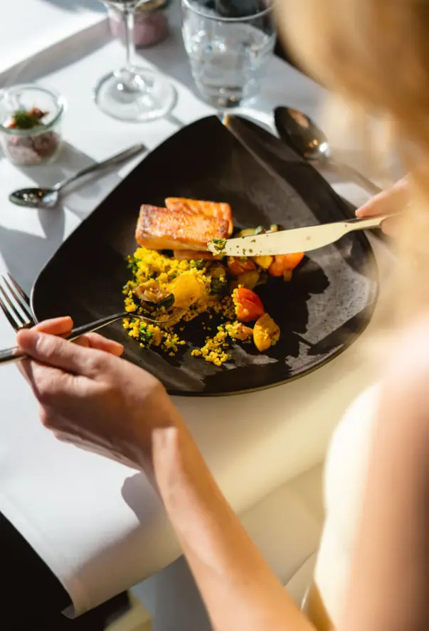 Eine Frau isst eine Mahlzeit in einem stilvollen Restaurant. Sie hält Messer und Gabel in ihren Händen, während sie ein Lachs-Quinoa Gericht von einem schwarzen Teller ist.  