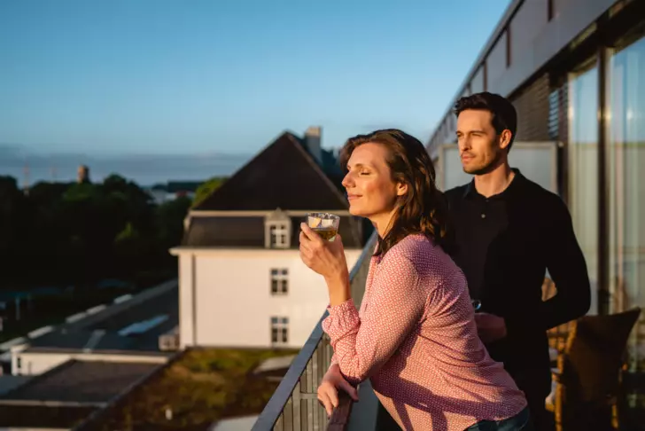 Ein Mann und eine Frau stehen gemeinsam auf einem Balkon, der eine Aussicht auf ein dämmriges Meer und Gebäude bietet. Die Frau, im Vordergrund, hält ein Glas Tee und hat die Augen geschlossen, während sie die Sonnenstrahlen genießt. Der Mann steht hinter ihr und blickt in die Ferne.