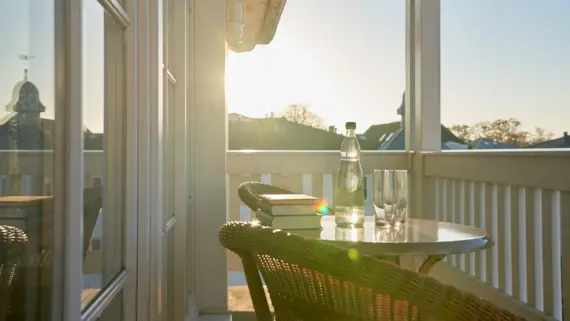 Ein einladender Balkon mit einem runden Tisch und zwei Korbstühlen, auf dem eine Flasche Wasser und Gläser im Sonnenlicht glänzen. Die warmen Sonnenstrahlen durchfluten die Szene und heben die entspannte Atmosphäre hervor. Im Hintergrund ist der blaue Himmel über den Dächern der Stadt zu sehen.