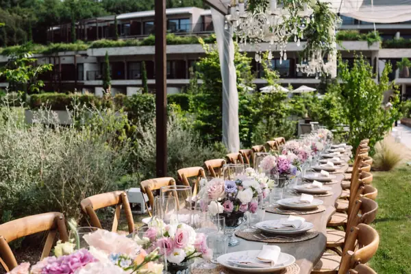 Lange Hochzeitstafel im Garten, dekoriert mit rosa und weißen Blumen, Holzstühlen und Blick ins Grüne.