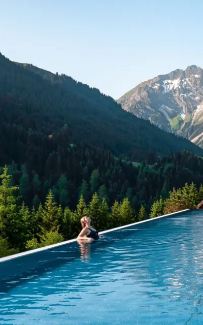 Frau lehnt am Rand eines Infinity Pools mit Bergkulisse im Hintergrund.