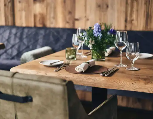 Ein rustikaler Holztisch gedeckt mit Weingläsern und Servietten sowie Blumendekoration und einer gepolsterten Sitzbank vor einer hölzernen Wand im Hindergrund..