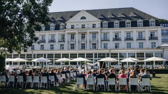 Eine Hochzeitszeremonie im Freien vor einem prachtvollen Hotelgebäude, bei der die Gäste auf weißen Stühlen sitzen und auf das Brautpaar blicken, das im Vordergrund steht. Elegante Sonnenschirme spenden Schatten auf der grünen Wiese, während das historische Kurhaus im Hintergrund eine klassische Kulisse bietet.
