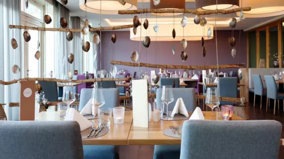 Modern eingerichtetes Dünenrestaurant im A-ROSA Hotel auf Sylt. Der Speisesaal ist hell und einladend, mit großen Fenstern, die viel Tageslicht hereinlassen. Hölzerne Dekorationselemente und hängende Muscheln verleihen eine maritime Atmosphäre, während die aufgeräumten Tische mit Wassergläsern, Weingläsern und Besteck für die Gäste vorbereitet sind.