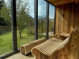 Holzbank in einem Raum mit Fenstern, durch die man ins Grüne, den Himmel und Berge im Hintergrund sehen kann.