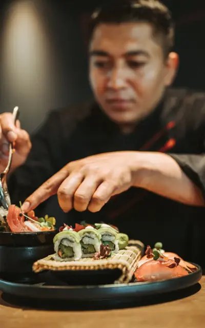 Ein Koch bereitet sorgfältig Sushi zu und legt die letzten Details an. Im Vordergrund sind verschiedene Sushi-Rollen und Nigiri auf einem Teller zu sehen. 