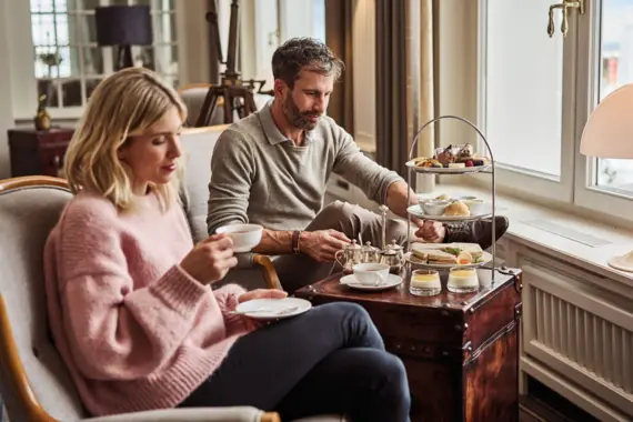 Ein Paar genießt eine stilvolle Tea-Time in einem eleganten Ambiente. Auf einem kleinen Tisch steht eine Etagere mit feinem Gebäck, Sandwiches und süßen Leckereien, begleitet von Tee und kleinen Desserts.