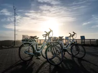 Das Foto zeigt mehrere weiße Fahrräder, die in der Sonne auf einem gepflasterten Weg geparkt sind. Im Hintergrund ist der Himmel klar und von sanften Wolken durchzogen. Die Sonne steht tief und bildet eine leuchtende Silhouette um die Fahrräder. Die Szene strahlt Ruhe und die Freude an der Natur aus.