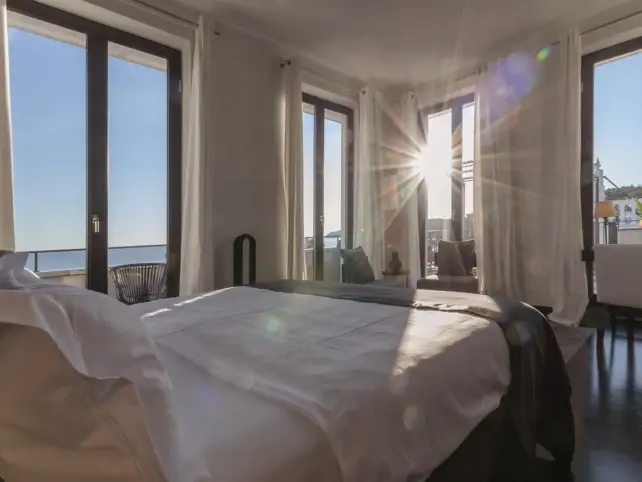 Ein aus einer Ecke fotografiertes Hotelzimmer mit gemütlichem Bett und bodentiefen Fenstern, durch die die Sonne hereinscheint und das Meer zu sehen ist. 