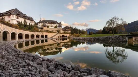  Das Bild zeigt eine beeindruckende Ansicht des A-ROSA Hotels in Kitzbühel während der Herbstzeit. Man sieht ein großes Gebäude im traditionellen alpenländischen Stil, das sich malerisch in die umgebende Landschaft mit ihren sanften Hügeln und dem bewaldeten Hintergrund einfügt. Im Vordergrund reflektiert ein ruhiger Teich die Architektur und die umgebenden Bäume, während eine Brücke mit Bogenkonstruktionen den Teich überquert und den Weg zum Resort weist. Die sanften Farben des Herbstes und das weiche Licht der untergehenden Sonne verleihen dem Foto eine ruhige und einladende Atmosphäre.