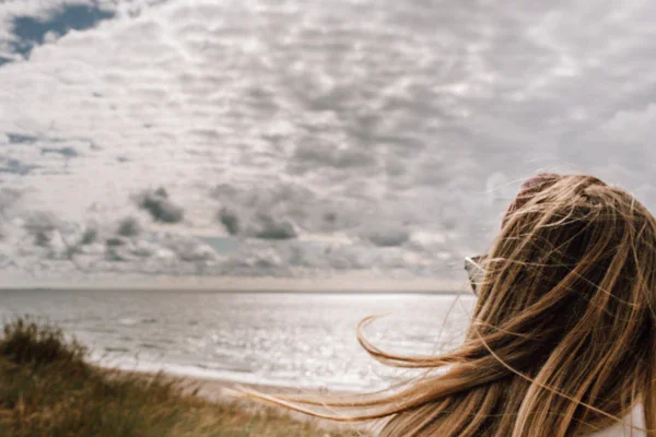 Eine Frau schaut auf die Weiten des vor ihr liegenden Meeres während die Sonne warm auf das Wasser scheint und ihre Haare im Wind wehen. 