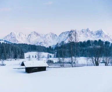 Eine verschneite Landschaft mit einer schneebedeckten Hütte im Vordergrund. Im Hintergrund sind viele Bäume und eine große Gebirgskette zu sehen.