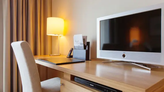 Ein heller Stuhl steht vor einem Schreibtisch auf dem ein kleiner Fernseher, ein Telefon sowie einige Flyer stehen.