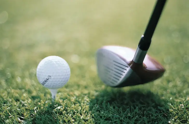 Auf einem grünen Goldplatz ist ein weißer Golfball platziert, welcher mit einem Golfschläger anvisiert ist. 