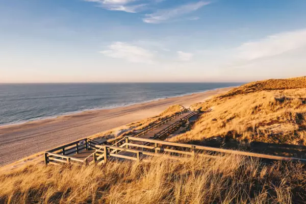 Ein Steg eingebettet in die Sylter Dünenlandschaft vor dem weißen Sandstrand und der Nordsee vor blauem Himmel im Hintergrund