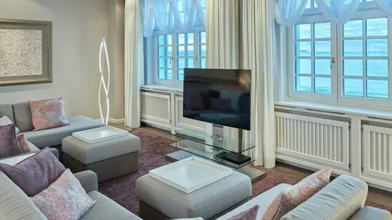 Ein elegantes Wohnzimmer in einer Suite mit Blick auf das Meer. Die Einrichtung besteht aus grauen Sofas und Hockern mit farblich abgestimmten Kissen. Im Zentrum steht ein moderner Couchtisch aus Glas und Weiß, über dem ein Fernseher platziert ist. Eine kreative Stehlampe fügt eine Designnote hinzu, während die klassischen weißen Vorhänge den Blick auf traditionelle weiße Fensterrahmen mit Aussicht auf die Ostsee freigeben. Über einem der Sofas hängt ein abstraktes Kunstwerk, das die schlichte Eleganz des Raumes unterstreicht.