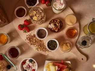 Tisch mit verschiedenen Frühstückskomponenten für ein Müsli, in kleinen Schüsseln auf einem Holzbrett.