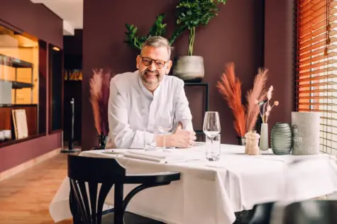 An einem Restauranttisch sitzt ein Koch in weißer Kochkleidung und lächelt freundlich in die Kamera. 