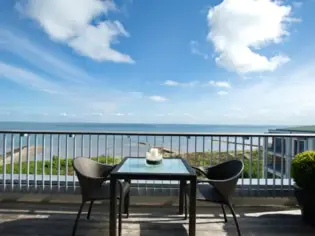 Eine Terrasse mit einem Tisch und zwei Stühlen, die vor dem Geländer stehen. Im Hintergrund ist das Meer und ein strahlend blauer Himmel zu sehen.