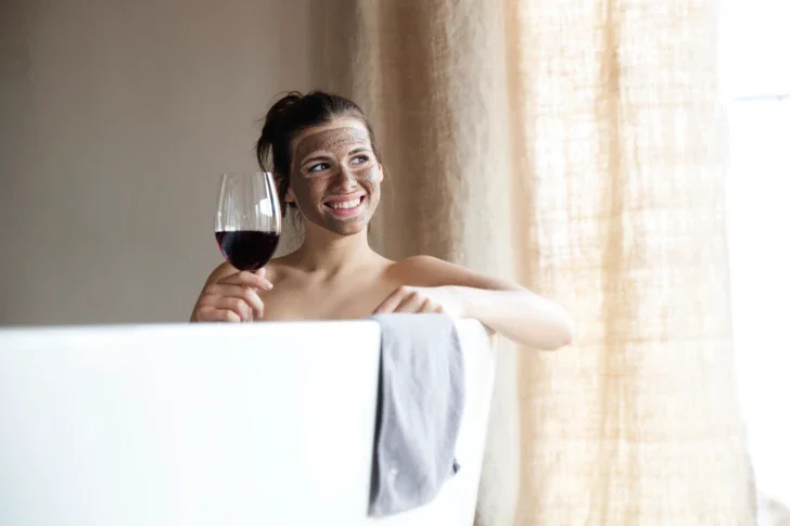 Eine Frau hält ein Glas Rotwein in ihrer Hand während sie mit einer Gesichtsmaske auf ihrem Gesicht lächelnd in einer weißen Badewanne sitzt.