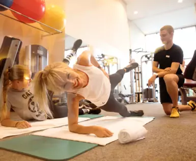 Eine Frau macht lächelnd Sportübungen auf einer Yogamatte, daneben knieten Personal Trainer.  