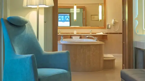 Ein Ausschnitt einer Suite mit einem eleganten blauen Sessel im Vordergrund und einem Blick auf das angrenzende Badezimmer. Im Badezimmer dominiert eine große Badewanne mit Marmorverkleidung, umgeben von sanftem Licht und einer entspannenden Atmosphäre. An der Wand sind moderne Leuchten angebracht, und auf einem Hocker liegt ein frisches Handtuch bereit. 