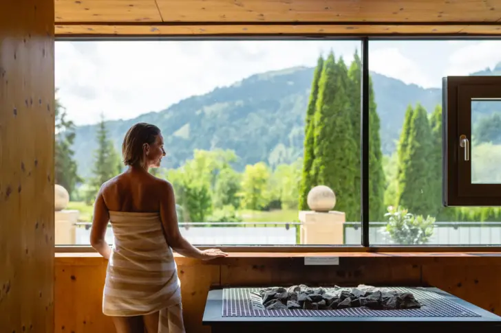 Eine Frau in einer Sauna, eingewickelt in ein weißes Handtuch, steht entspannt vor einem großen Fenster und blickt auf eine malerische Berglandschaft und einen Garten. Sie steht neben einem Saunaofen. 