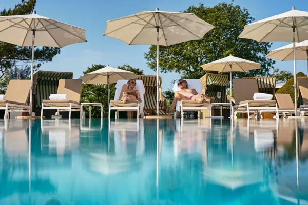 Mann lehnt am Rand eines Infinity-Pools mit Blick auf einen Ort inmitten einer grünen Berglandschaft im Hintergrund.