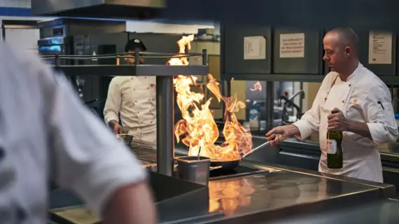 Ein Koch bereitet ein Gericht in einer Pfanne mit Flammen zu. Um ihn herum stehen weitere Köche. 