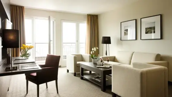 A bright living area with a beige sofa and two armchairs. On the left is a dark wood desk and a terrace can be seen in the background.