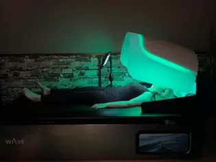  Eine Frau entspannt sich auf einem modernen Wellnessgerät, dem Wellsystem Wave, einer Liege, die mit einer leuchtenden grünen Kuppel über dem Kopfteil ausgestattet ist. 