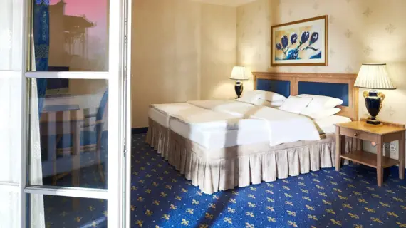 Ein elegant gestaltetes Doppelzimmer in einem Hotel, das durch seine warmen Farbtöne und die klassische Einrichtung Gemütlichkeit ausstrahlt. Das große Bett mit pristiner weißer Bettwäsche und einem gesteppten Kopfende in sanften Blautönen ist das Herzstück des Raumes. Darüber hängt ein farbenfrohes Gemälde, das farblich auf die blauen Akzente im Raum abgestimmt ist. Die Nachttische sind mit stilvollen Lampen versehen, deren sanftes Licht eine entspannte Atmosphäre schafft. Der Boden ist mit einem blauen Teppich bedeckt, der kleine gelbe, florale Motive aufweist. Eine geöffnete Fenstertür mit weißen Sprossen lässt die Verbindung zu einem Balkon erahnen.