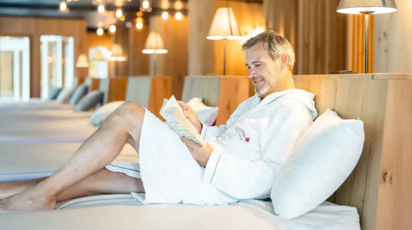 Mann in weißem Bademantel sitzt auf einem Bett und liest ein Buch.