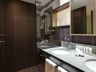 A bathroom with dark tiles, a shower and a vanity unit with two washbasins.