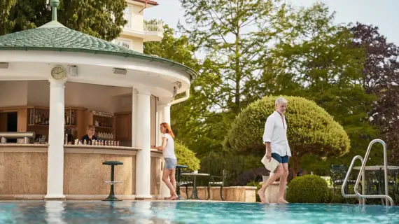 Außenpool mit Blick auf die stilvolle Poolbar im A-ROSA Strandidyll Heringsdorf, eine Frau bestellt ein Getränk, ein Mann läuft entspannt am Poolrand entlang.