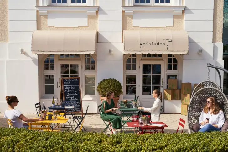 Eine Aussenansicht einer sonnigen Terrasse vor einem Weinladen, auf der Menschen an bunten Tischen und Stühlen sitzen und Wein trinken. 