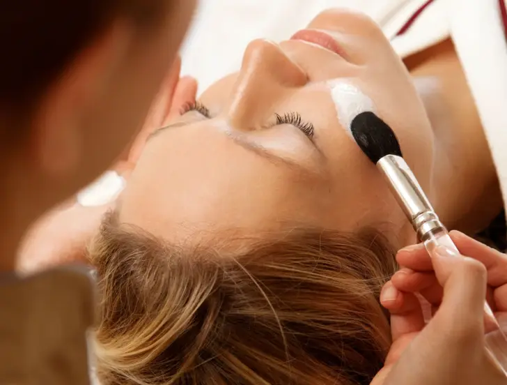  Eine Frau genießt eine entspannende Gesichtsbehandlung, bei der eine Kosmetikerin sorgfältig eine Gesichtsmaske mit einem Pinsel aufträgt. Die Frau liegt mit geschlossenen Augen da