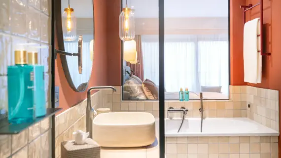 Modernes Badezimmer mit Waschbecken, Badewanne und Spiegel mit Blick auf ein Schlafzimmer, durch ein Fenster im Hintergrund..