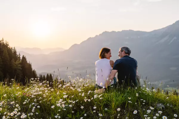 Ein Paar sitzt auf einer Blumenwiese in den Bergen. Im Hintergrund verläuft eine grüne Berglandschaft und der Himmel ist in warme sommerliche Farben getaucht. 