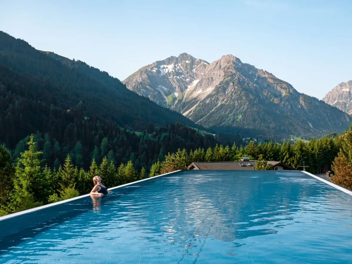 Eine Frau lehnt am Beckenrand in einem Infinity Pool unter freiem Himmel mit Bergen im Hintergrund.